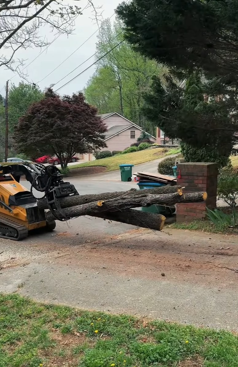 Heavy equipment loader carrying large cut tree logs out of a residential driveway